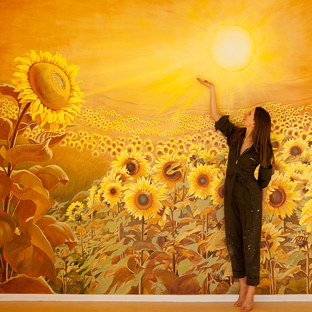 Elena Wuest vor dem Wandbild mit einem großen Sonnenblumenfeld und einer kräftig leuchtenden Sonne